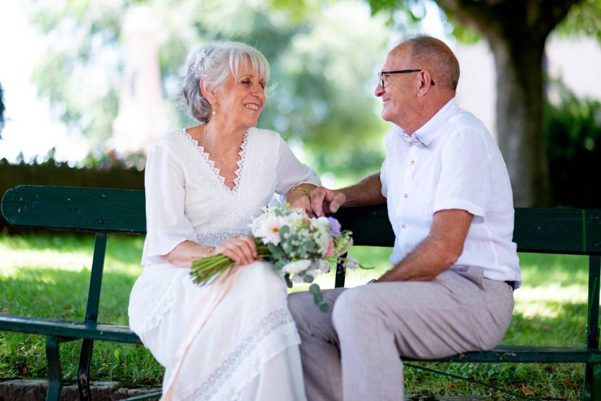 Couple âgé assis sur un banc dans un parc, échangeant un sourire complice. La femme en robe blanche tient un bouquet de fleurs, symbole de leurs 56 ans de mariage et des noces de lapis-lazuli, célébrant un amour fidèle et serein.